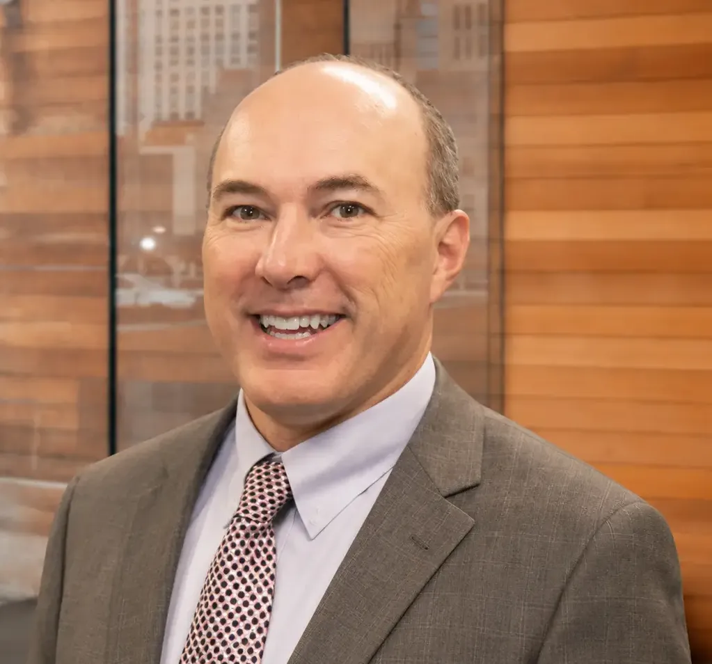 Professional man smiling in a business suit with a patterned tie, standing in front of a wooden wall backdrop, conveying a friendly and approachable demeanor in a corporate setting.